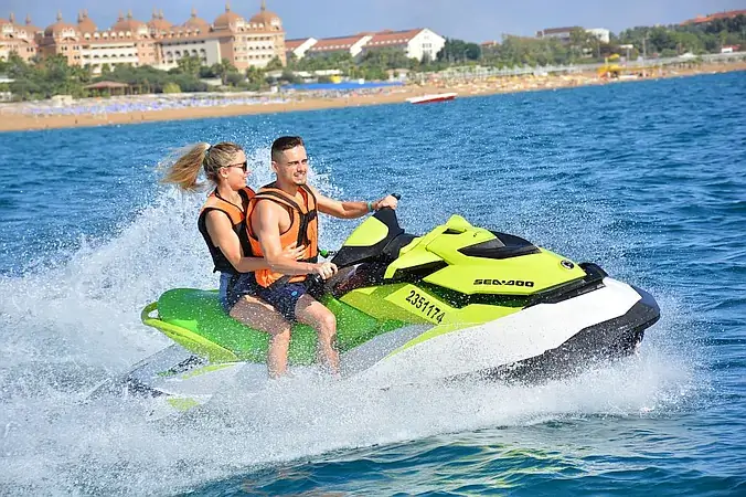 A thrilling Jet Skiing ride along the coastline — a man drives while a woman holds him, surrounded by splashing waves and open water.